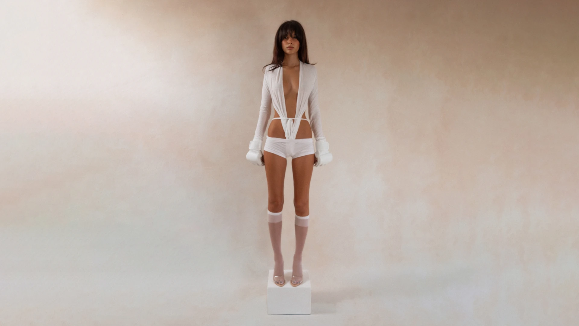 Model standing on a white block in a white plunging top, blush backdrop